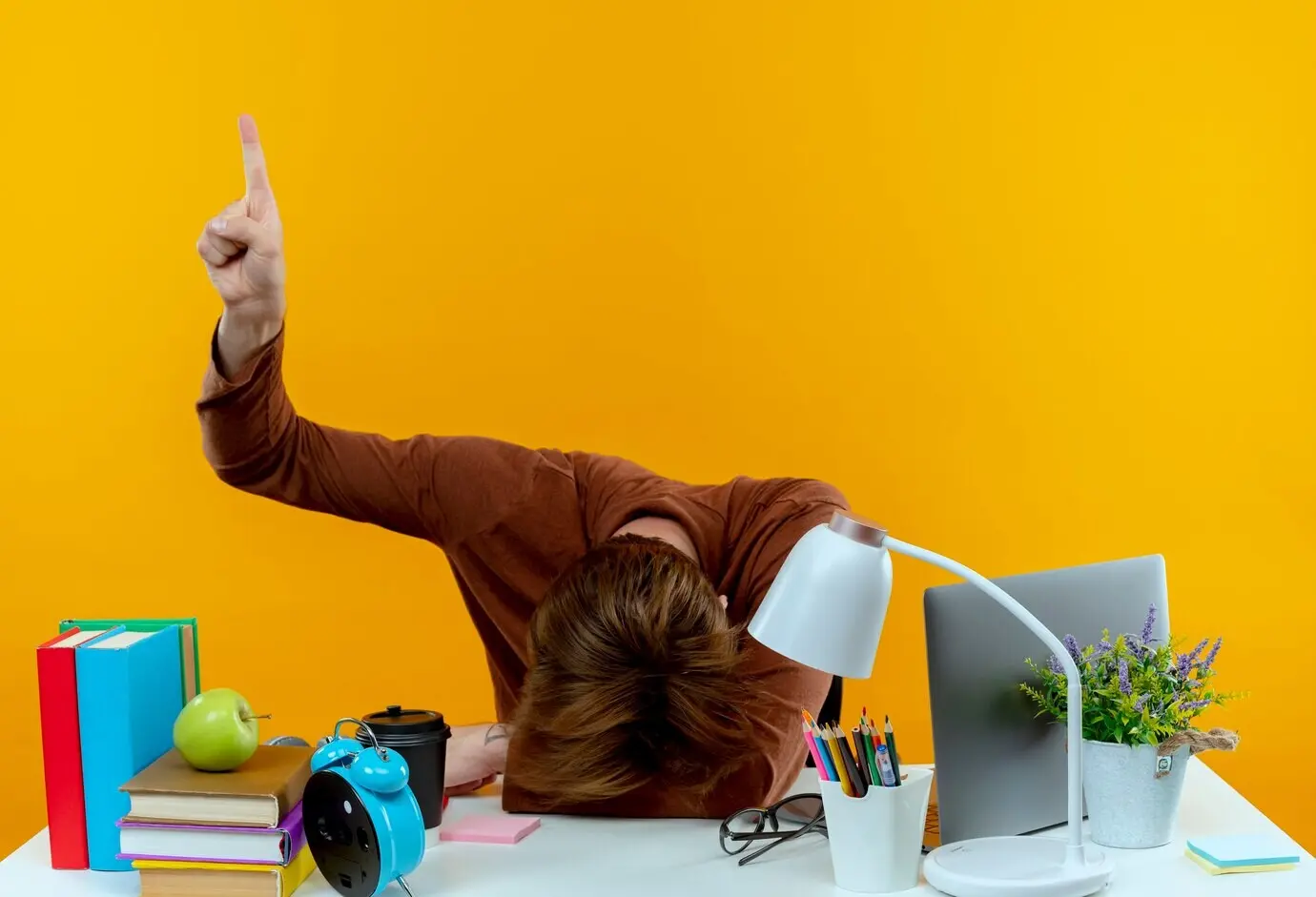 A young schoolboy with his head lowered sits at a desk with school supplies, pointing upward, isolated against a yellow wall with copy space.