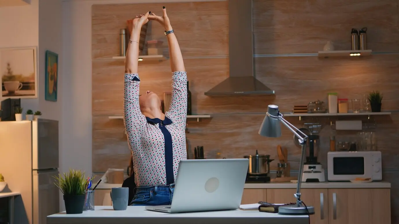 A tired woman working from home stretches after typing on a laptop late at night. A busy, focused employee using modern wireless network technology, working overtime for her job, reading, writing, and searching.